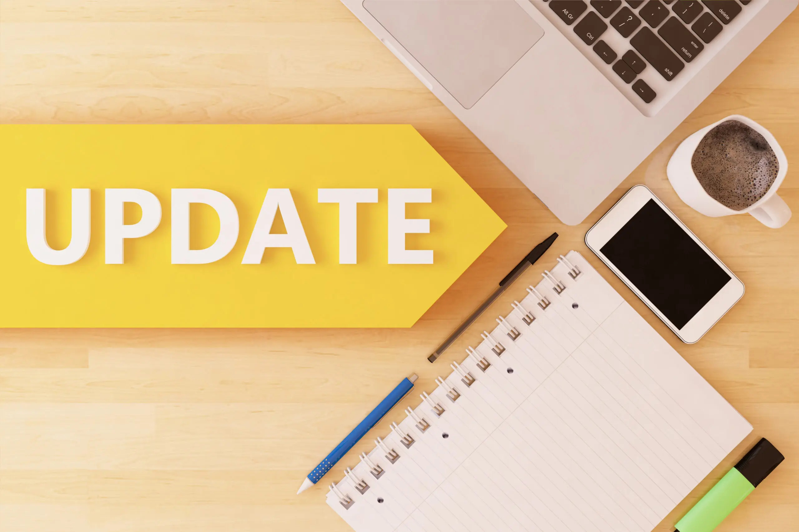 The word update written on a yellow tag banner, placed next to a Mac laptop, mobile phone, notebook, pens and coffee