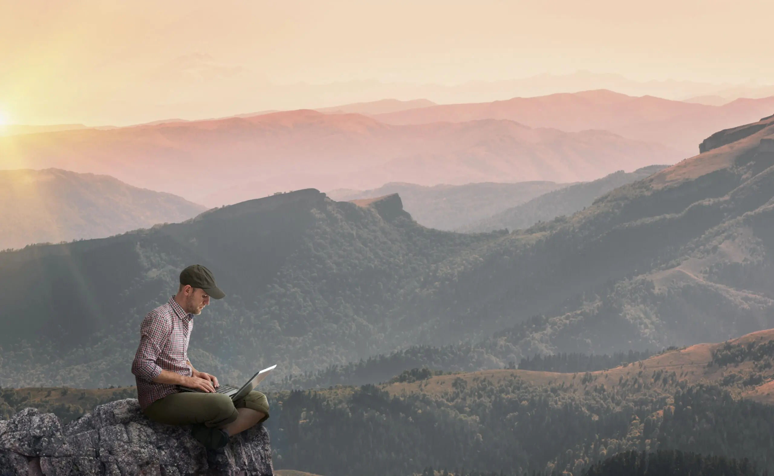 Large Image of a man sat on a cliff working on his Mac laptop