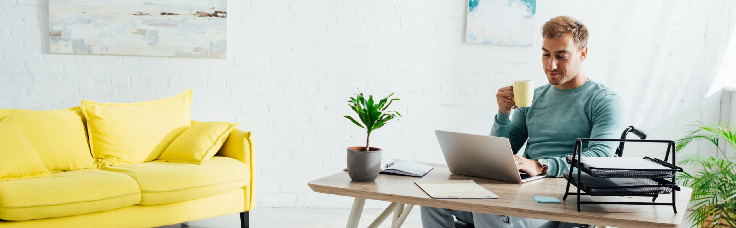 Young man sat at his home desk, working on his Mac laptop whilst drinking some coffee, with an open notebook