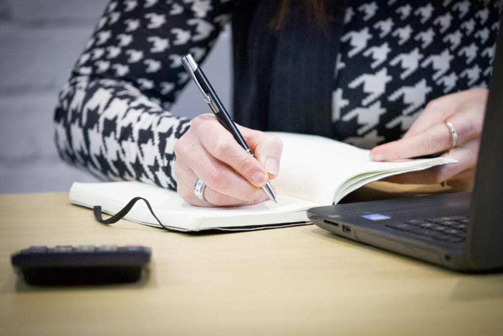 woman writing in her notebook with her laptop in front of her