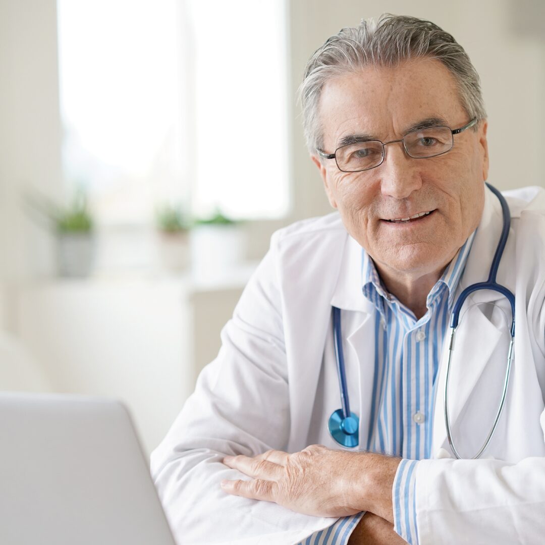 Smiling elder man in his doctors uniform, sat with his Mac laptop