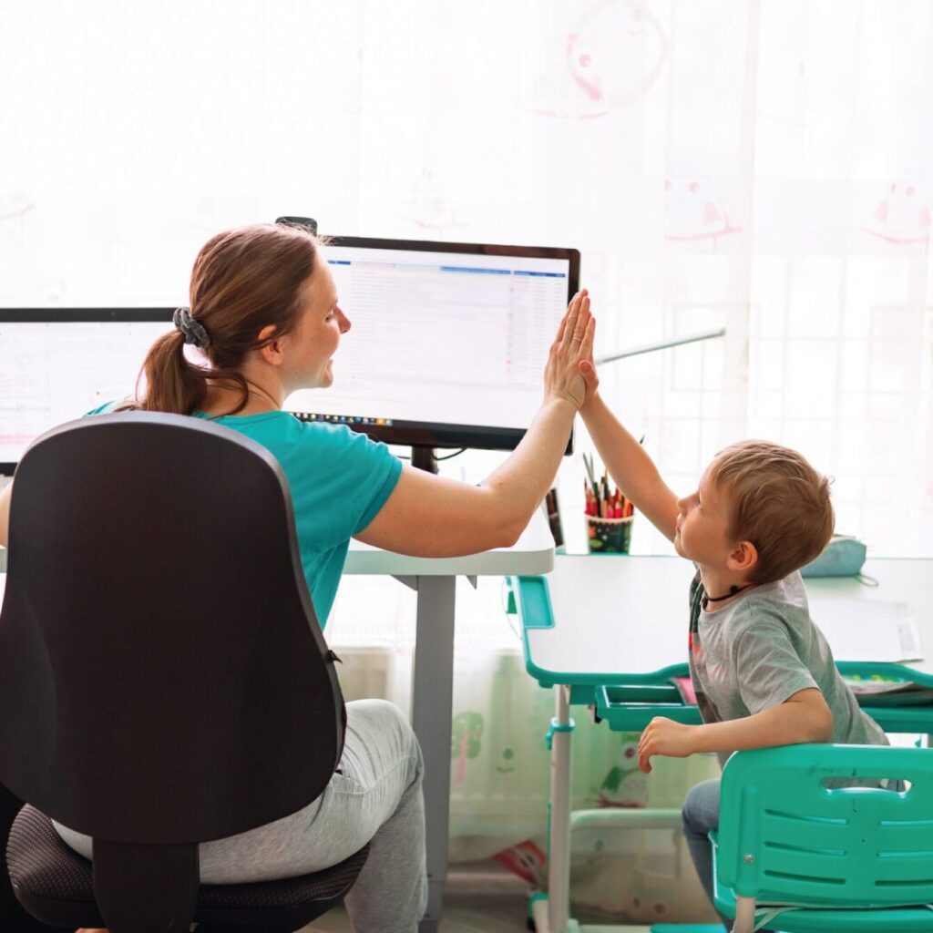 Lady working from her home desk, high-fiving her young son who is sat next to her on a kids desk