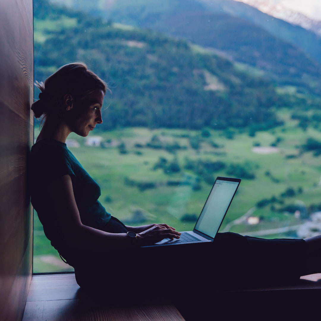 Woman working on her Mac laptop, with views of green hills and landscape behind her