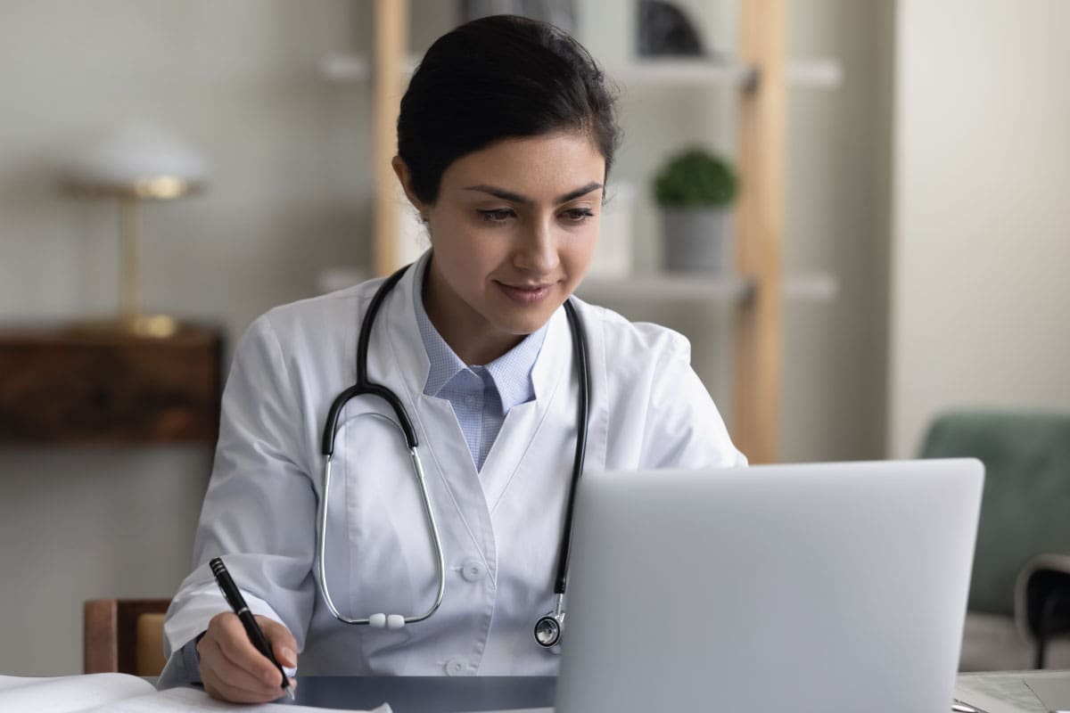 Young Nurse sat at her home desk, working on her Mac laptop whilst writing notes down