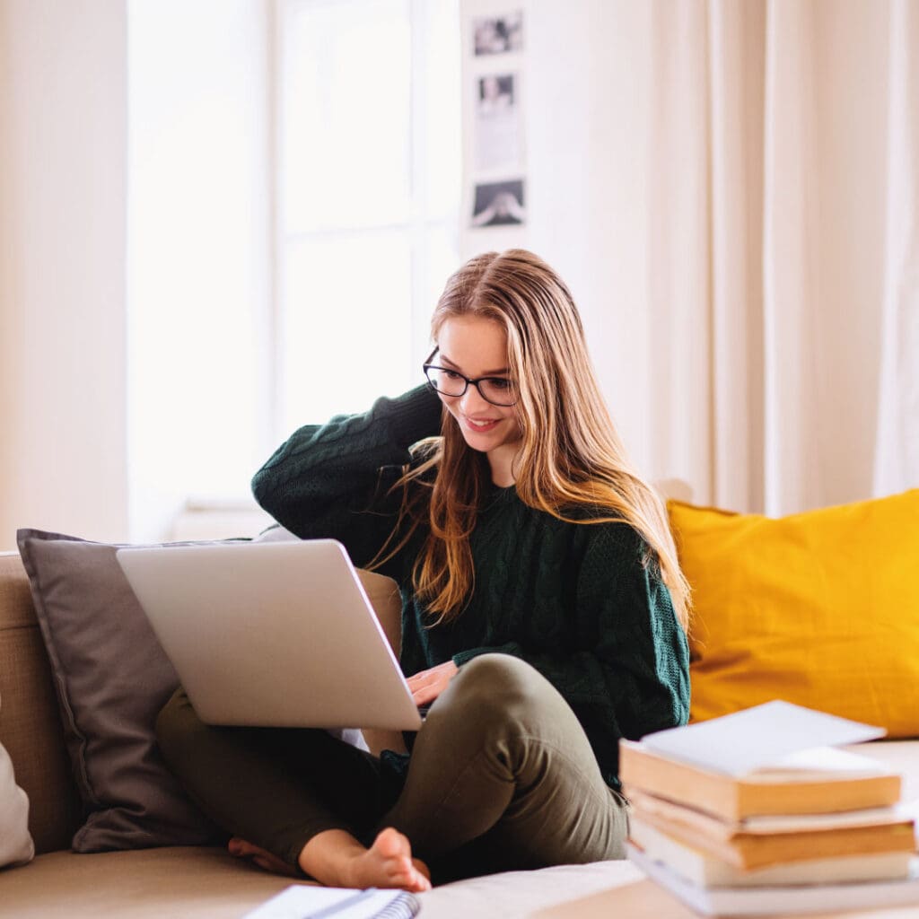 Young woman sat comfortably on her sofa, whilst working on her Mac laptop, with books placed next to her