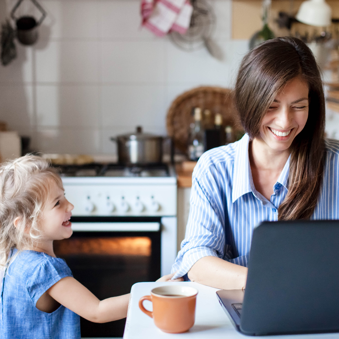 Woman sat at her kitchen table, working on her laptop whilst laughing and smiling with her young daughter besides her