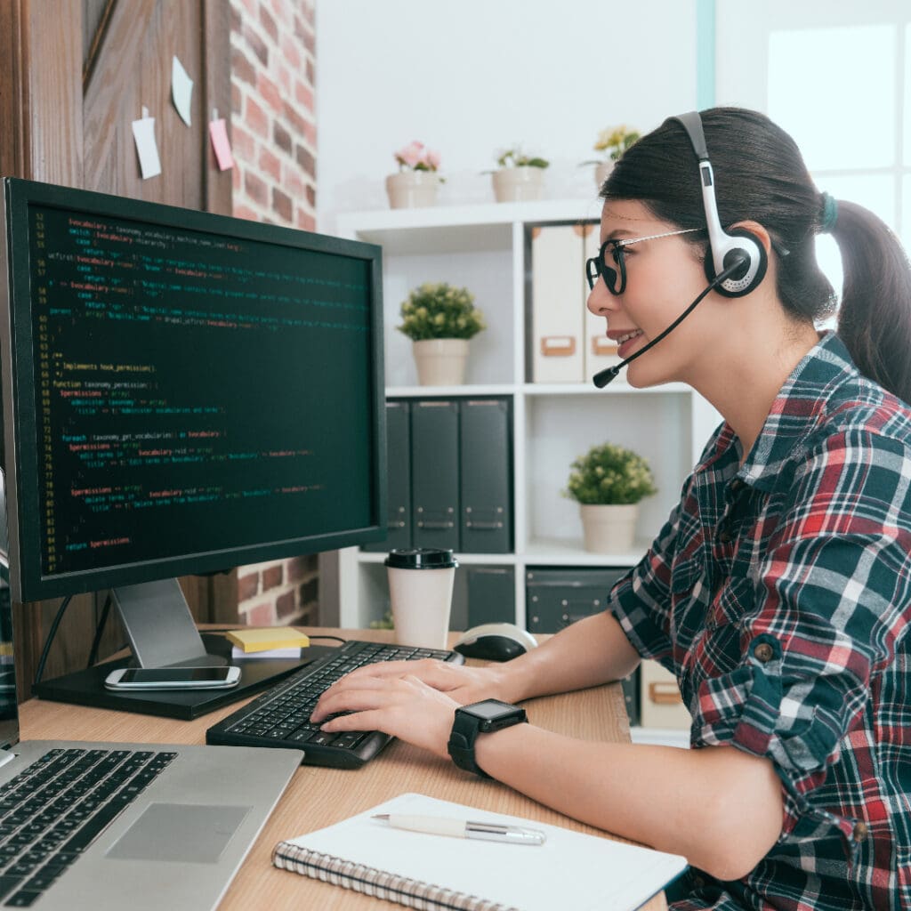 Young woman sat at her home desk with a headset on, working on a Mac laptop, as well as a monitor with coding on the screen