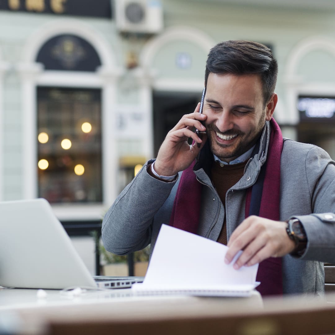 Smiling man working at a table outside, whilst on the phone, looking through a notebook with a Mac laptop besides him