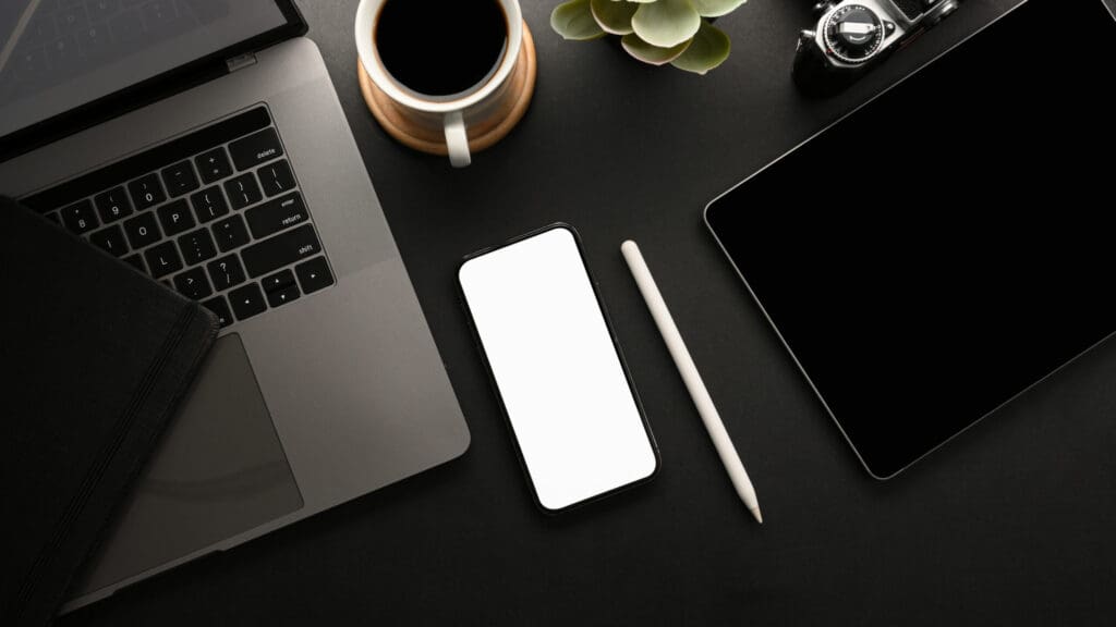 Laptop, iPad, iPhone, apple pencil and a notebook on a black desk