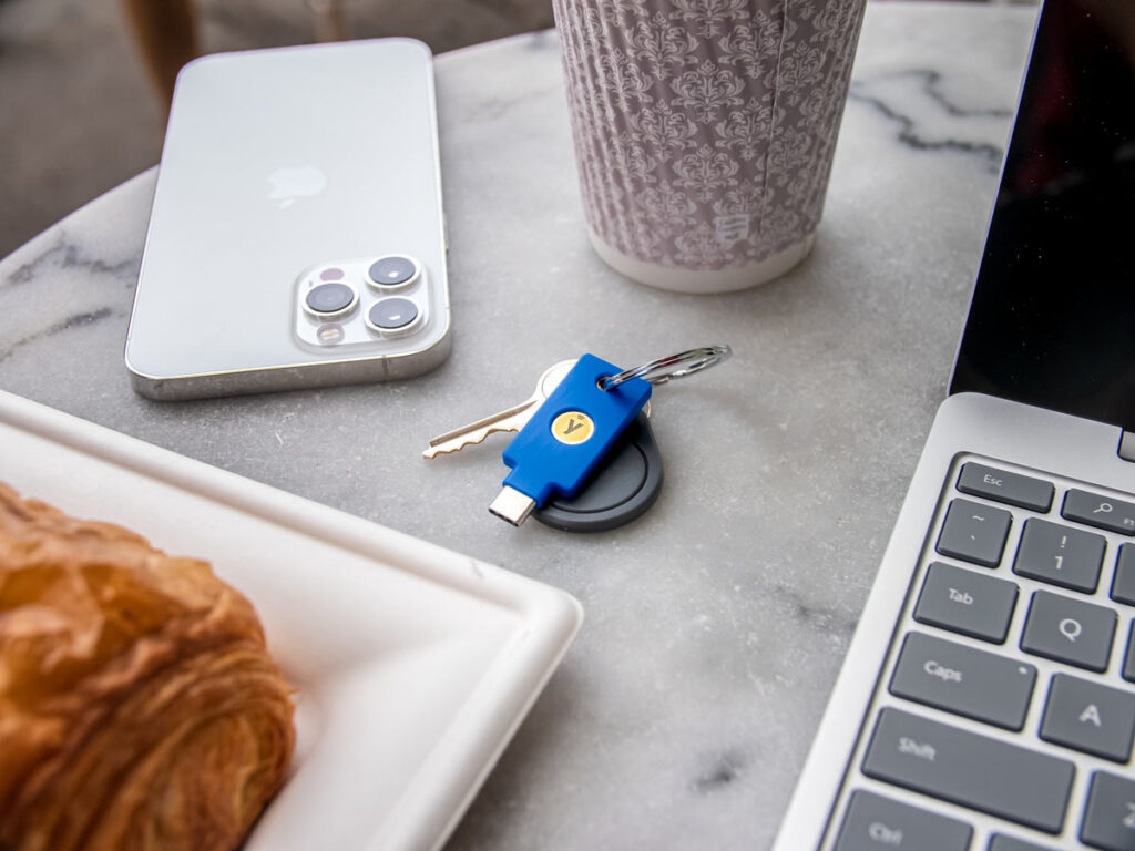 YubiKey security key C nfc on a keyring with a key and a fob, placed on table beside a laptop, mobile phone, coffee and pastry